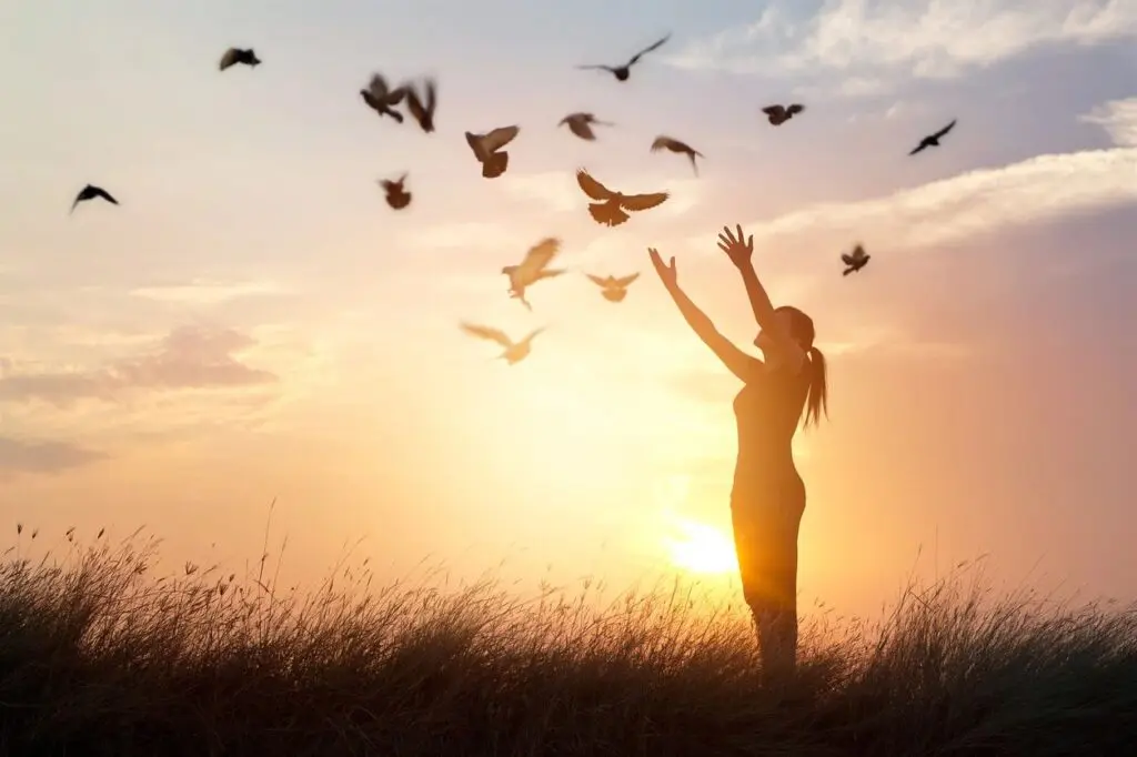 A woman standing in the grass with birds flying around her.